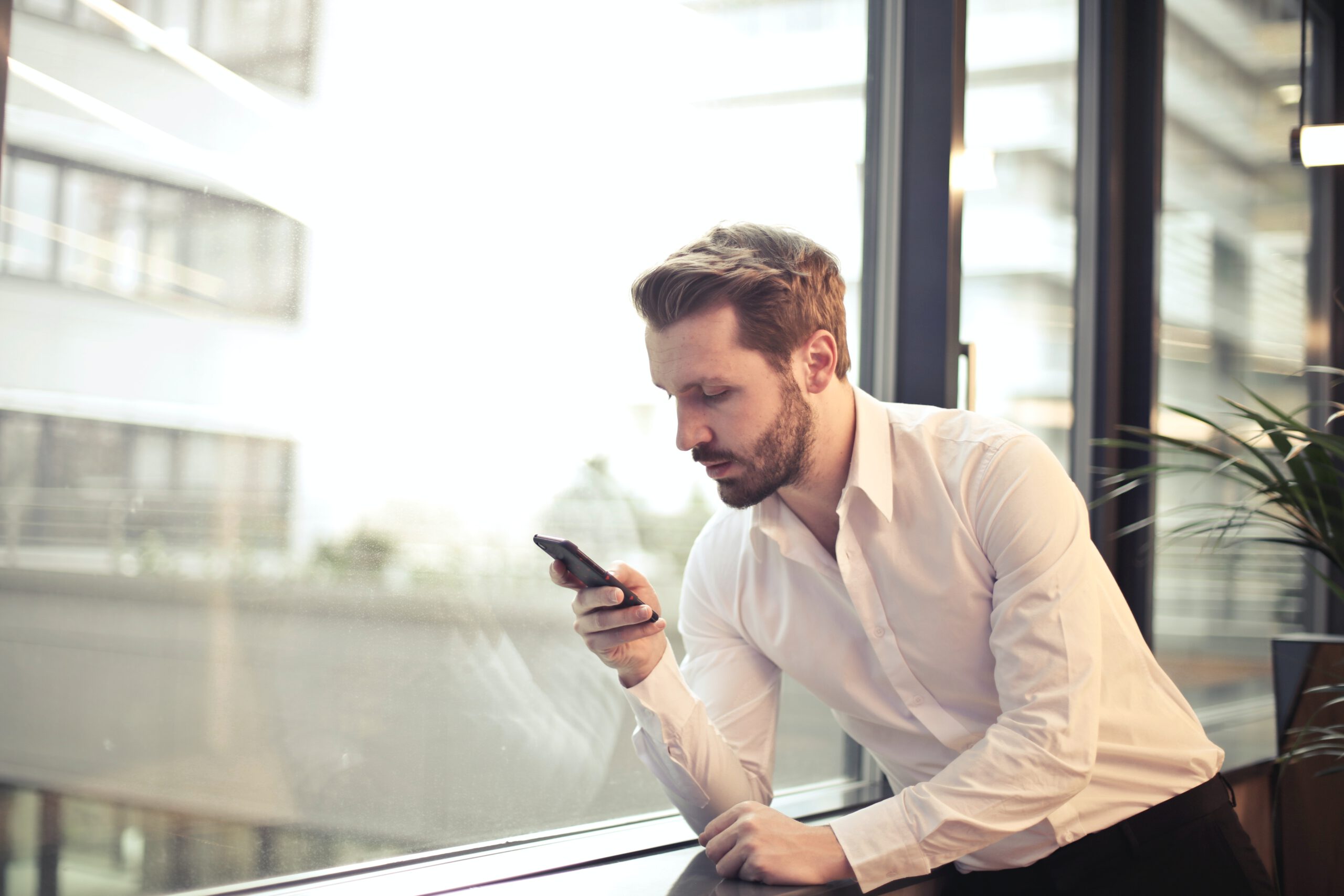 Mężczyzna w białej koszuli oparty o parapet i patrzący na ekran telefonu komórkowego