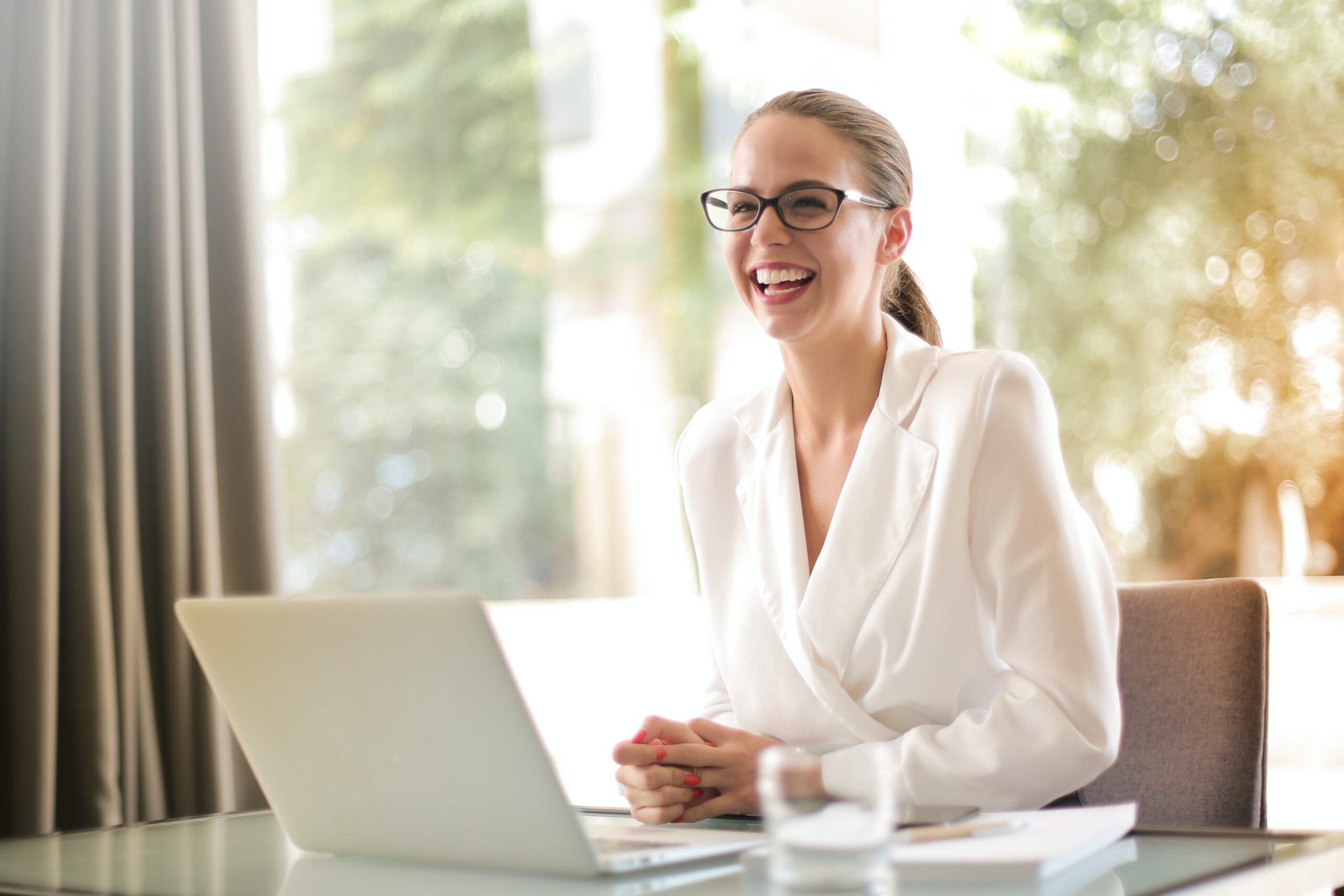 Śmiejąca się kobieta w okularach i białej koszuli, siedząca przy stole przed laptopem