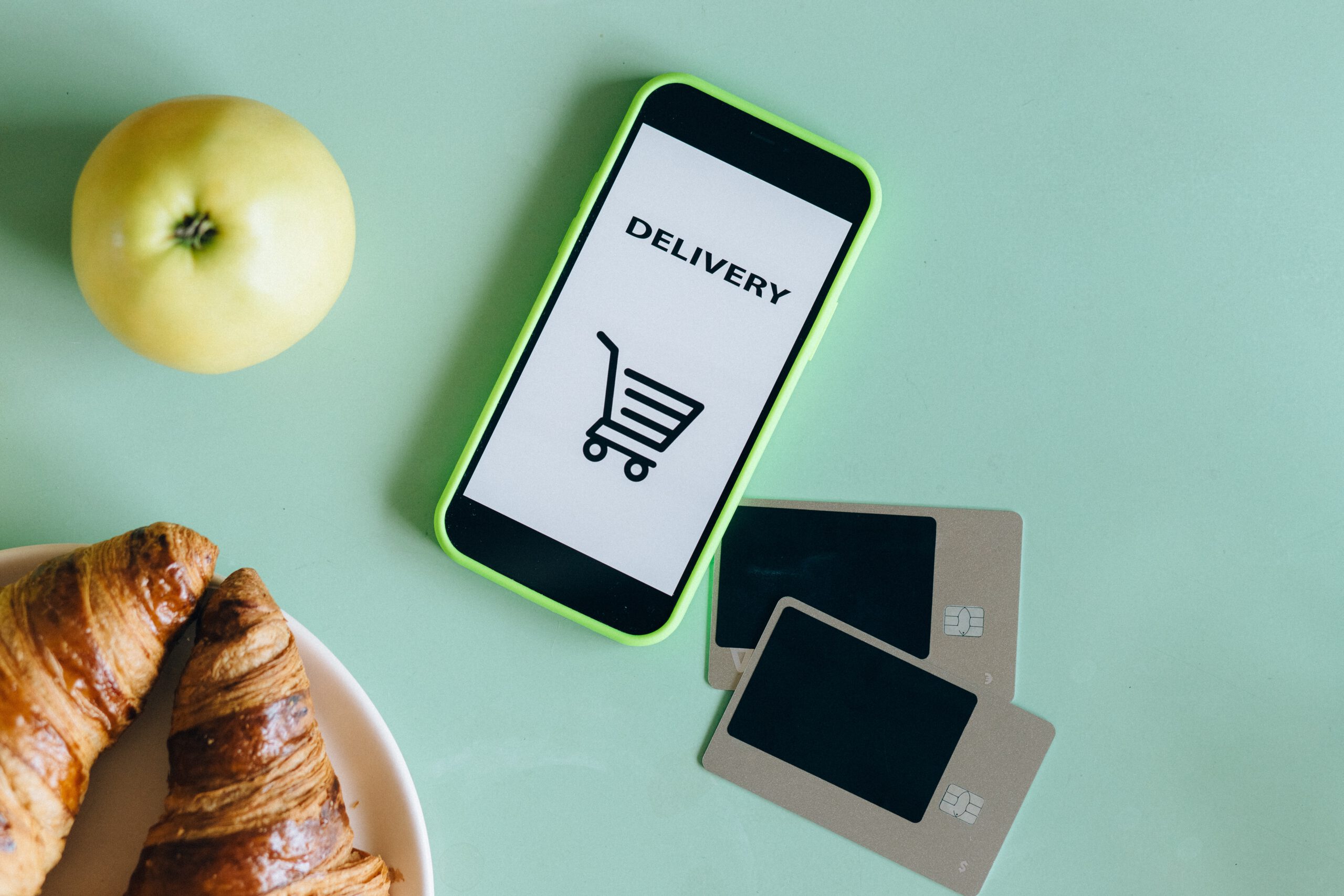 Telefon z wyświetloną grafiką wózka zakupowego, karty kredytowe i rogaliki, leżące na stole